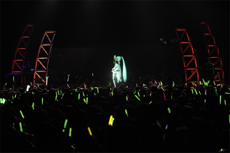 Hatsune Miku animated figure performing on a dark stage above green glow sticks