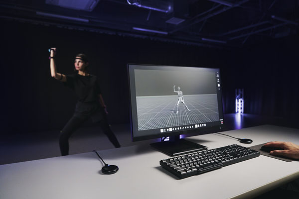 A performer wearing mocopi sensors beside a monitor showing a captured 3D figure